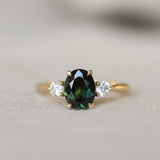 A yellow gold three-stone ring featuring an oval dark green sapphire in the center, flanked by two round clear accent stones, shown on a soft neutral background.