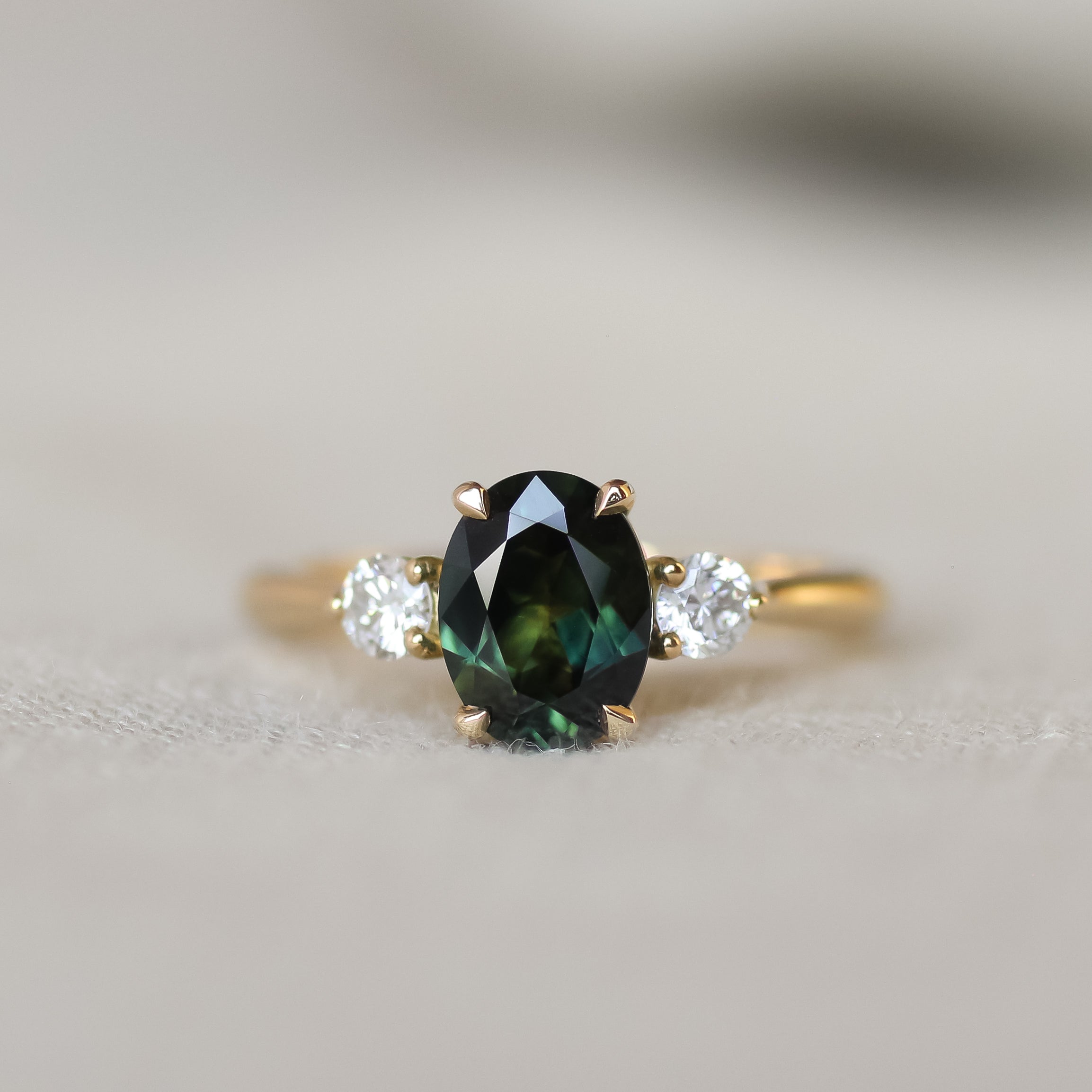 A yellow gold three-stone ring featuring an oval dark green sapphire in the center, flanked by two round clear accent stones, shown on a soft neutral background.