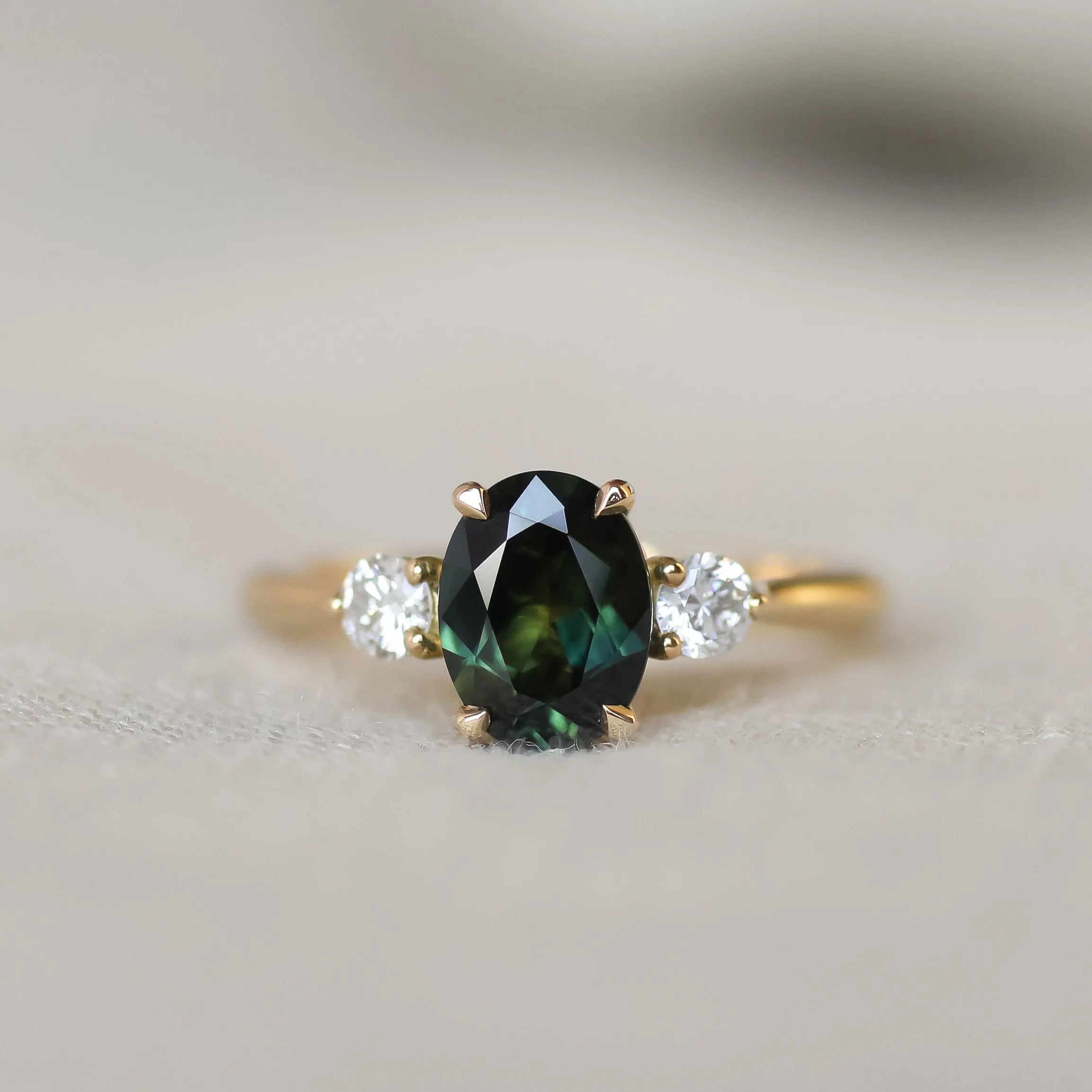 A yellow gold three-stone ring featuring an oval dark green sapphire in the center, flanked by two round clear accent stones, shown on a soft neutral background.