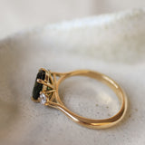 A gold ring with a dark green oval gemstone and a small accent diamonds, resting on a textured light beige stone surface.