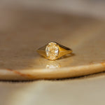 A gold signet ring with an oval-cut yellow diamond, resting on a polished stone slab with a soft-focus beige background.