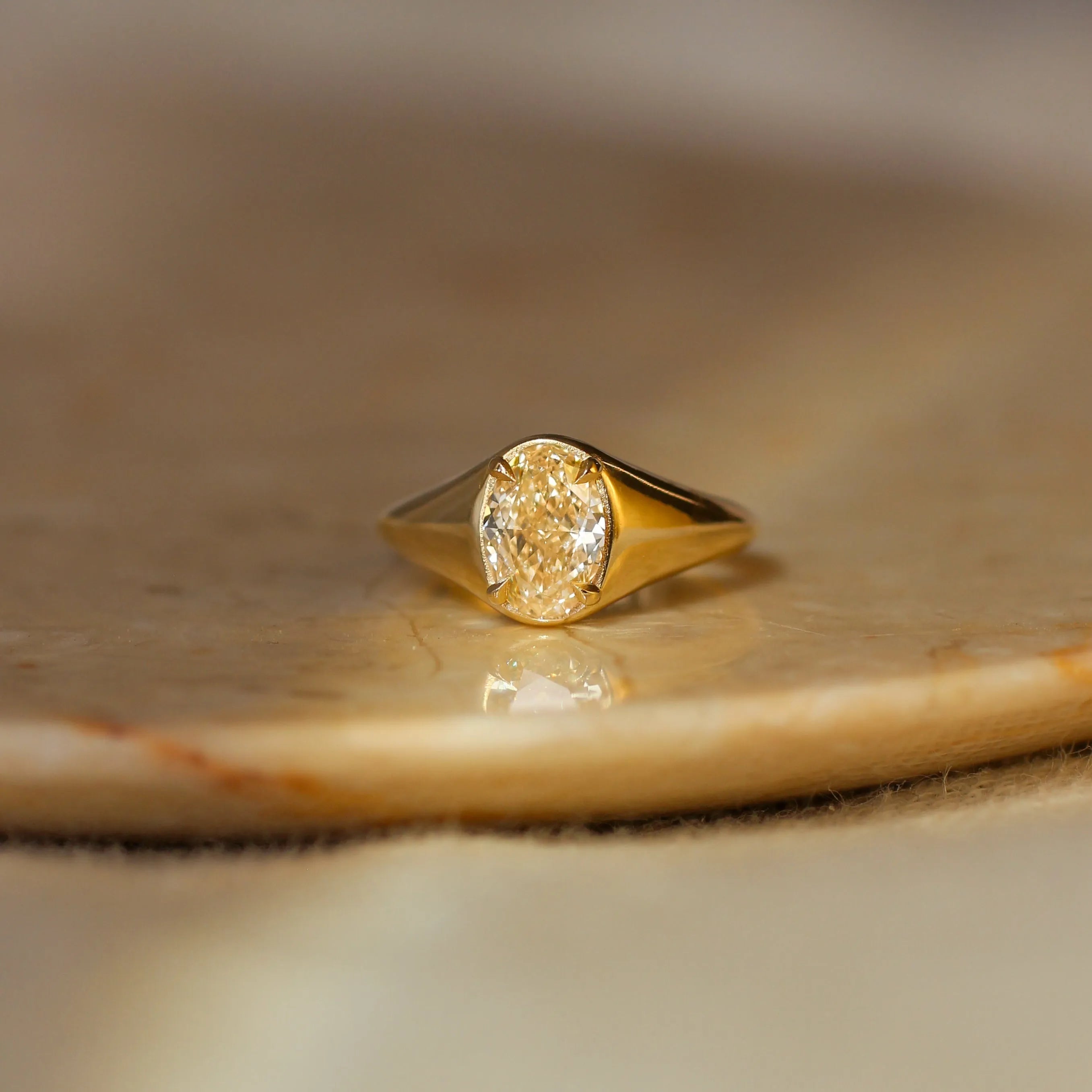 A gold signet ring with an oval-cut yellow diamond, resting on a polished stone slab with a soft-focus beige background.