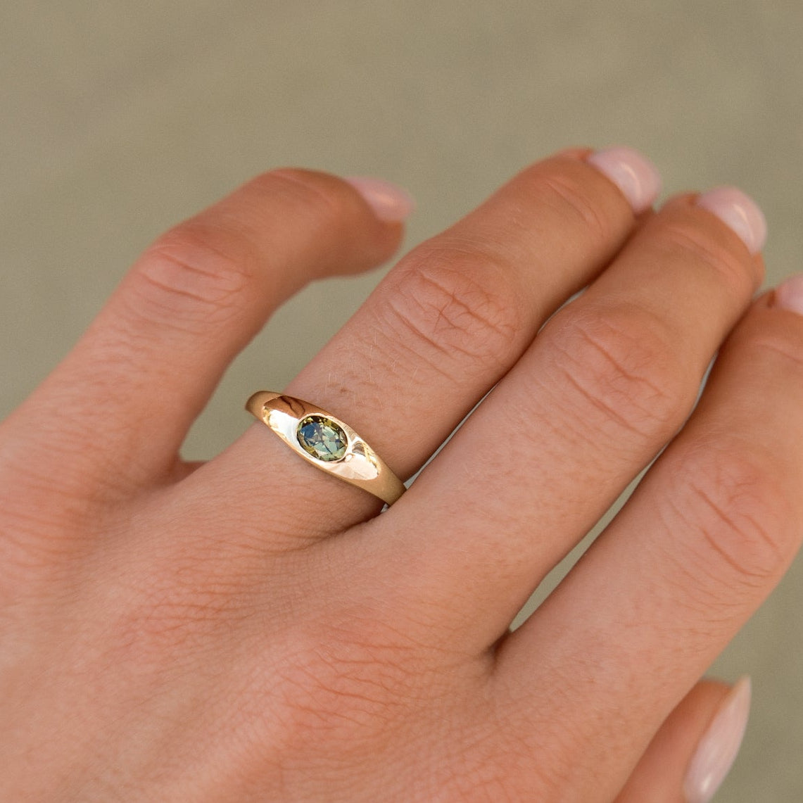 A close-up of a hand wearing a gold ring with a small oval green gemstone, set against a soft beige background.