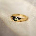 yellow gold band ring with a bezel set olive green oval sapphire in the centre, set on a beige fabric surface