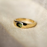yellow gold band ring with a bezel set olive green oval sapphire in the centre, set on a beige fabric surface