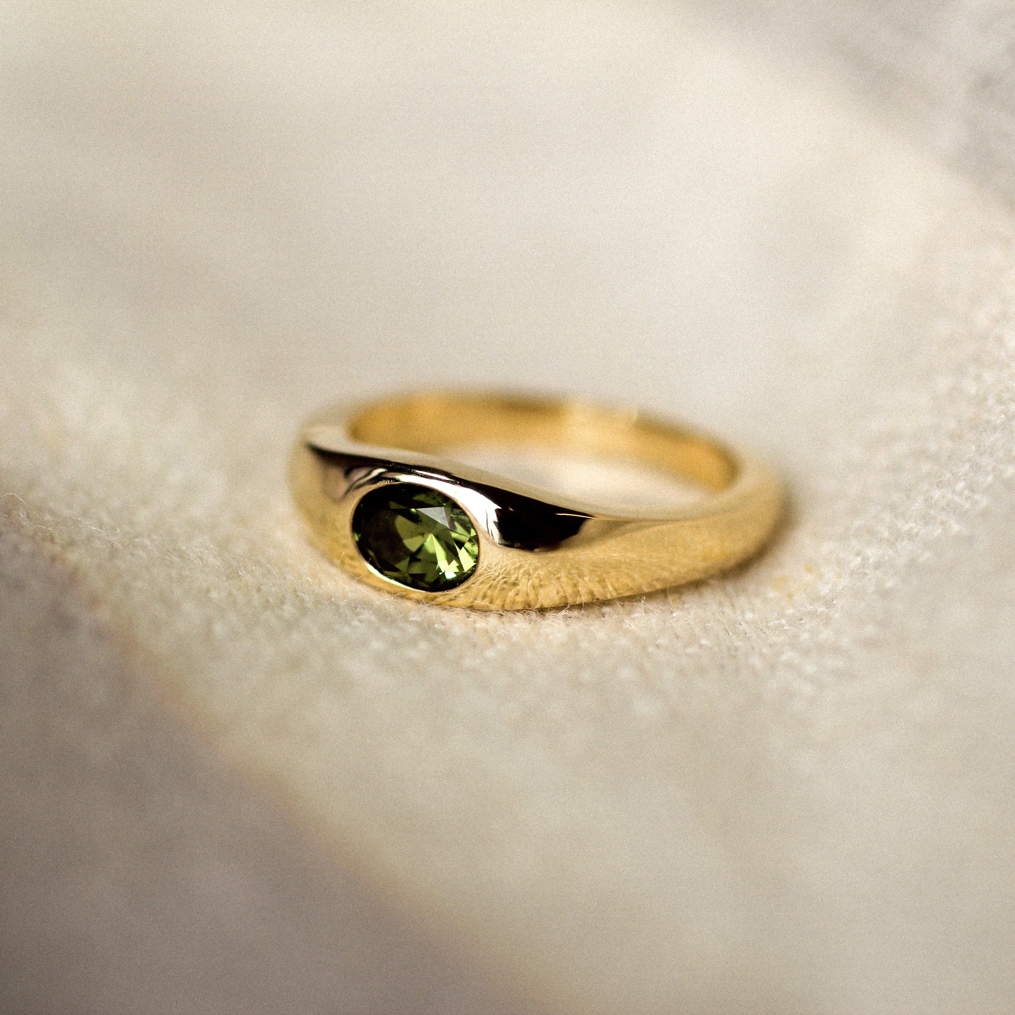 yellow gold band ring with a bezel set olive green oval sapphire in the centre, set on a beige fabric surface