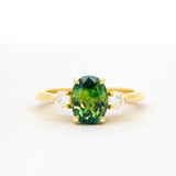A yellow gold ring featuring an oval-cut green gemstone in the center, flanked by two small clear round-cut stones on a white background.