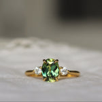 A gold ring featuring a central green oval gemstone and two small clear side stones, resting on a soft white textured surface with a blurred background.