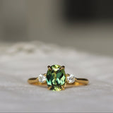 A gold ring featuring a central green oval gemstone and two small clear side stones, resting on a soft white textured surface with a blurred background.
