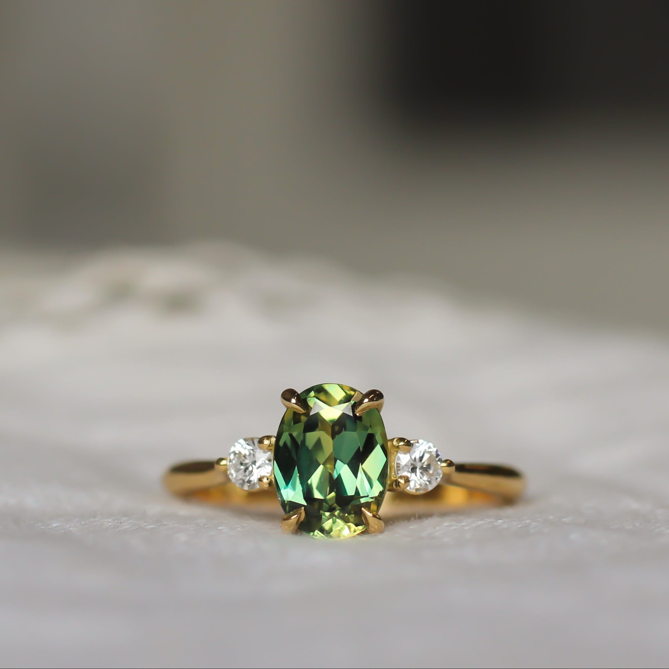 A gold ring featuring a central green oval gemstone and two small clear side stones, resting on a soft white textured surface with a blurred background.