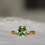 A gold ring featuring a central green oval gemstone and two small clear side stones, resting on a soft white textured surface with a blurred background.