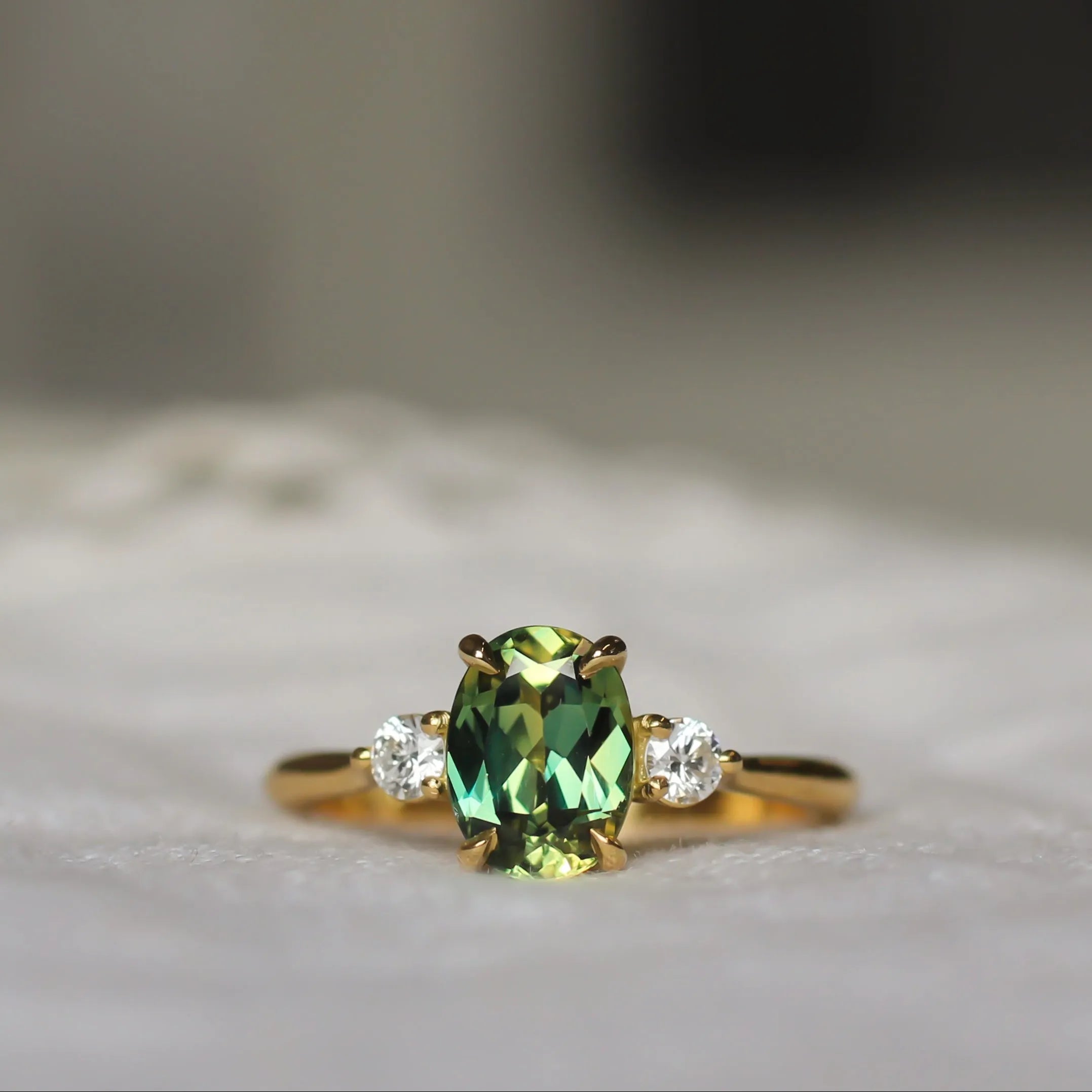 A gold ring featuring a central green oval gemstone and two small clear side stones, resting on a soft white textured surface with a blurred background.