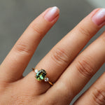 A close-up of a hand wearing a gold engagement ring with a green oval-cut sapphire and small diamond side stones, set against a soft gray background.
