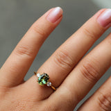A close-up of a hand wearing a gold engagement ring with a green oval-cut sapphire and small diamond side stones, set against a soft gray background.