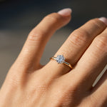 A close-up of a hand wearing a gold solitaire engagement ring with a round-cut clear gemstone, set against a softly blurred dark background.