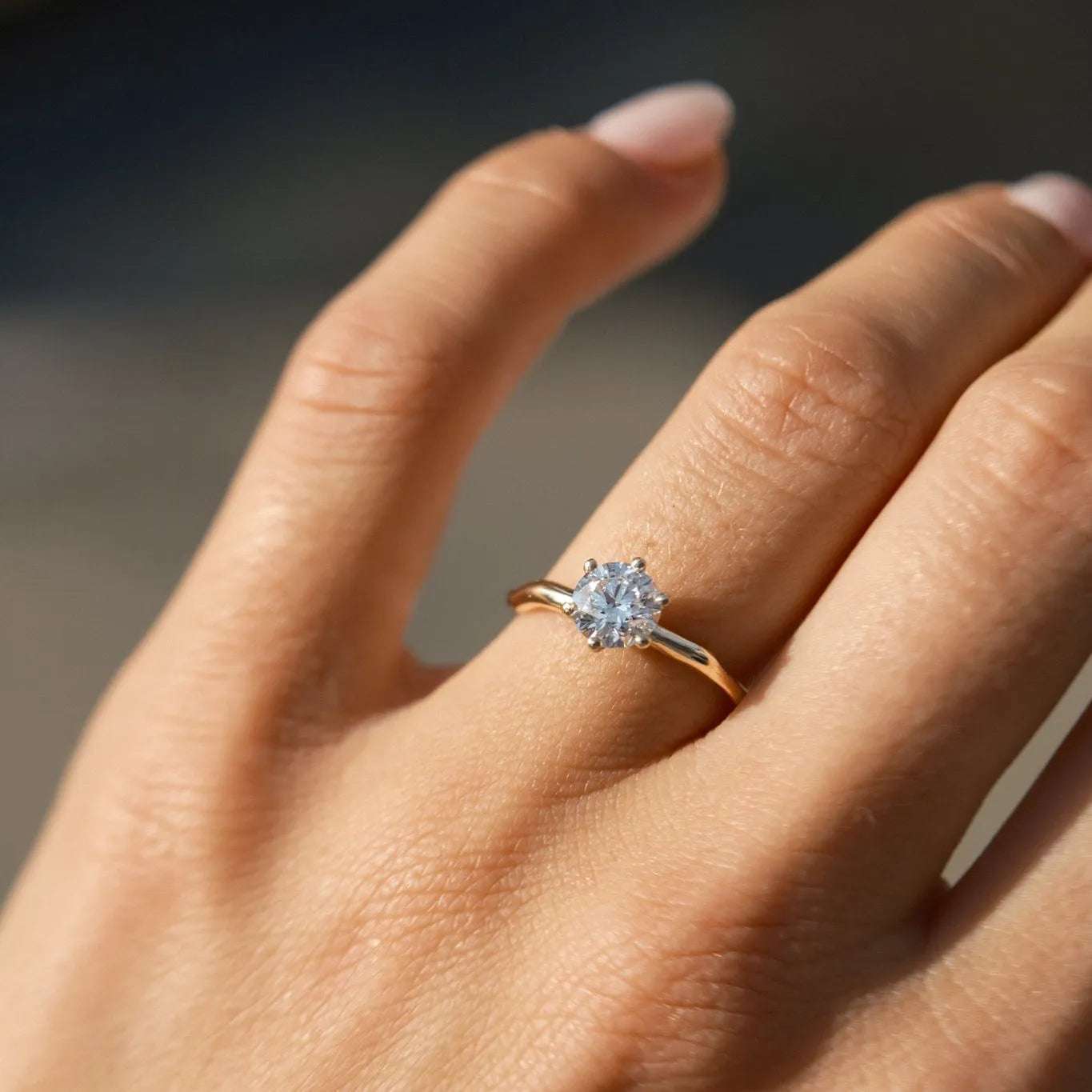 A close-up of a hand wearing a gold solitaire engagement ring with a round-cut clear gemstone, set against a softly blurred dark background.
