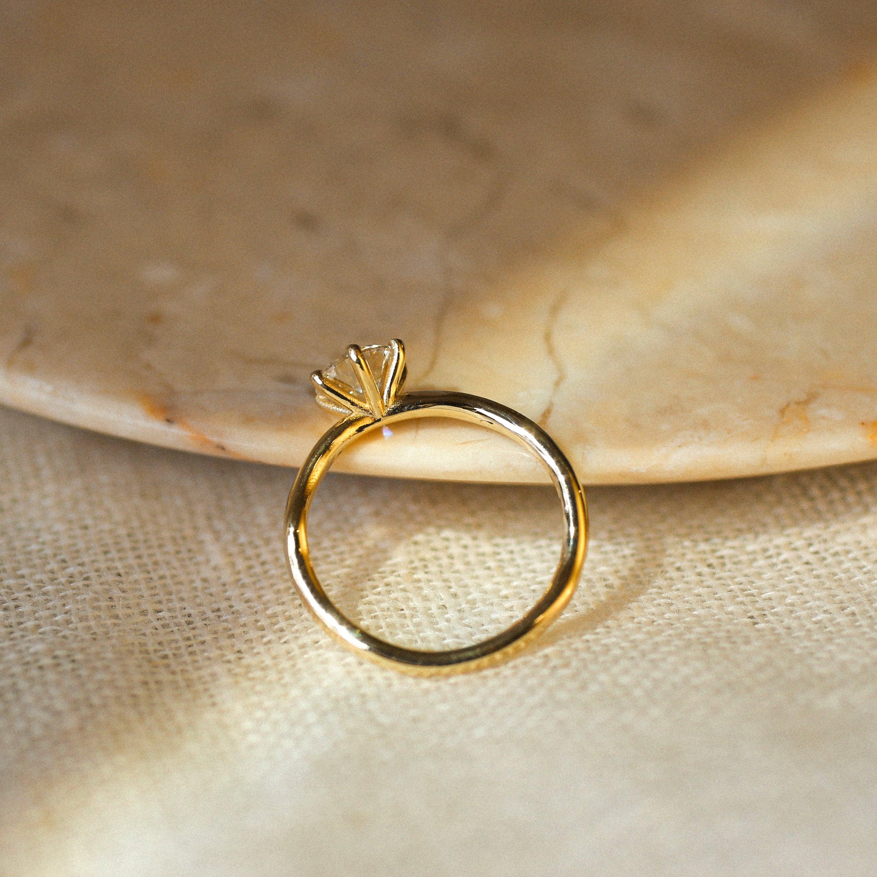 A gold ring with a high-set prong setting, photographed resting on a beige textured surface with a marble plate in the background.