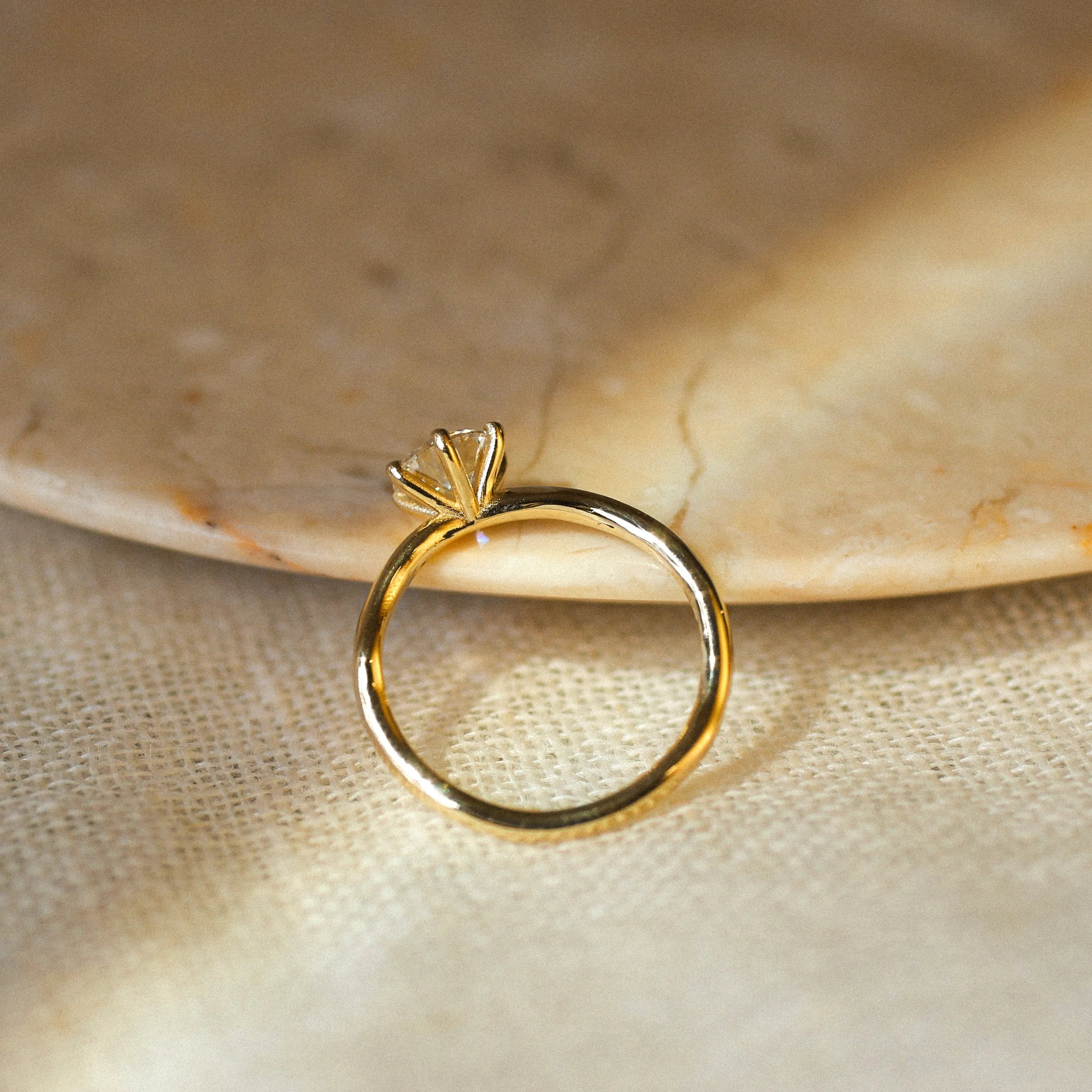 A gold ring with a high-set prong setting, photographed resting on a beige textured surface with a marble plate in the background.
