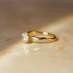 A yellow gold solitaire ring with a clear round-cut diamond and wavy molten organic band, resting on a warm beige marble surface under soft, directional sunlight.