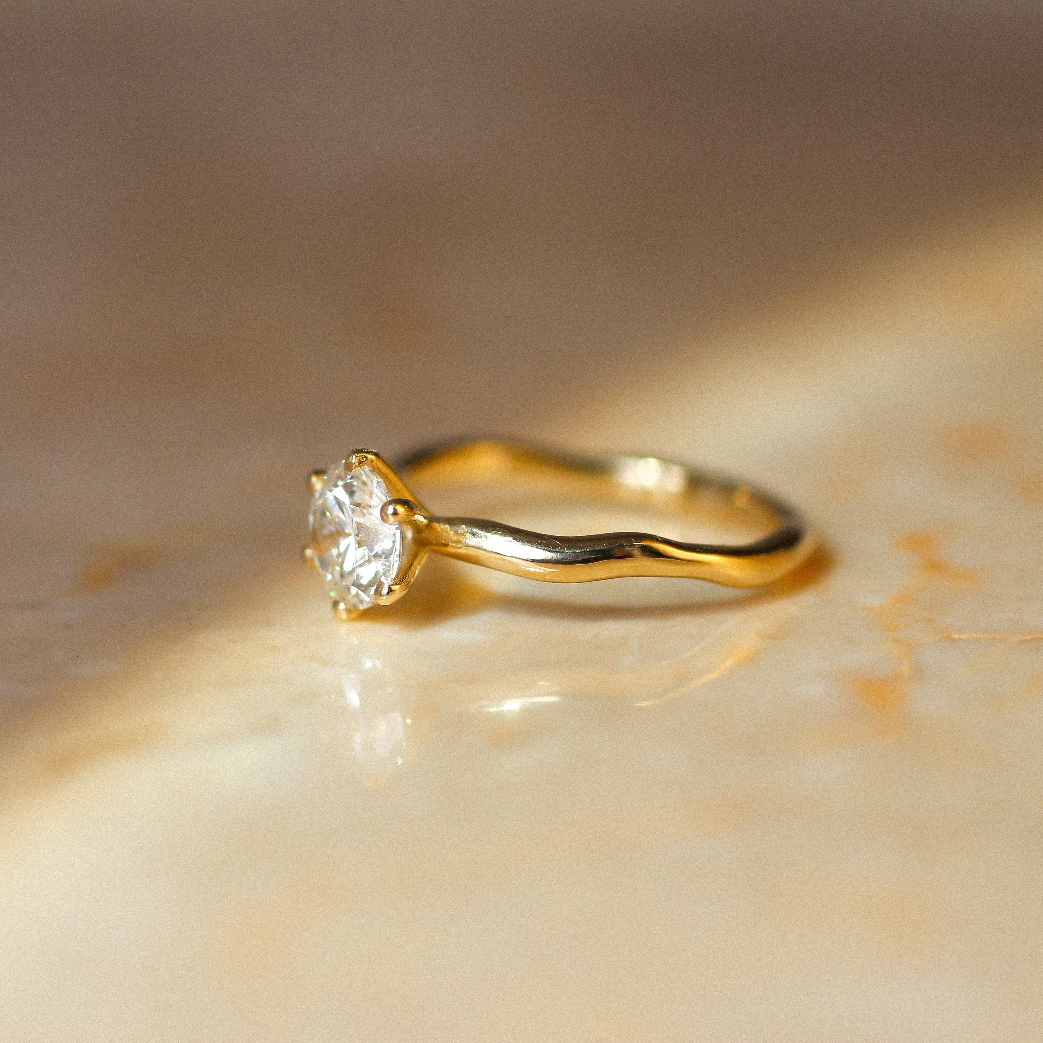 A yellow gold solitaire ring with a clear round-cut diamond and wavy molten organic band, resting on a warm beige marble surface under soft, directional sunlight.
