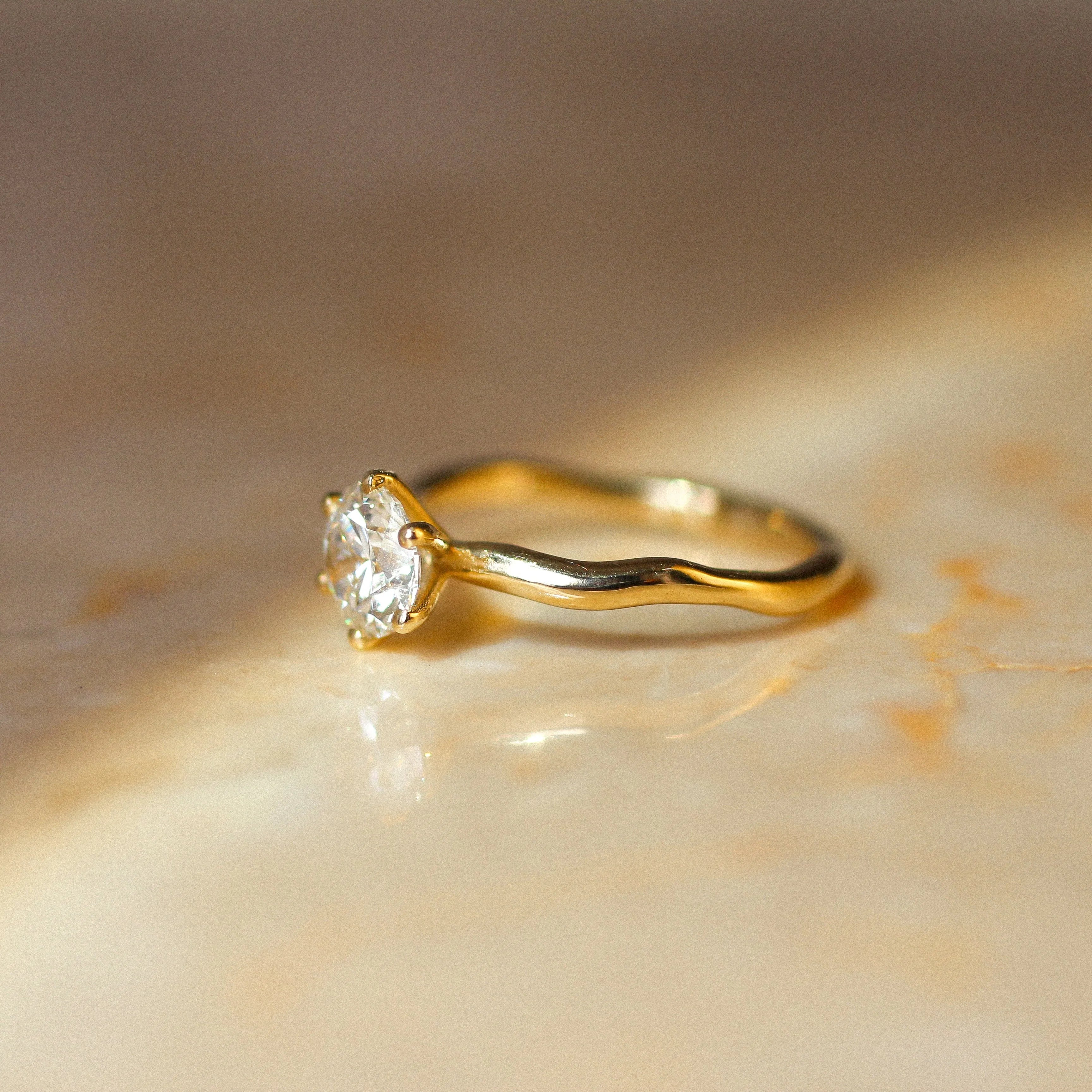 A yellow gold solitaire ring with a clear round-cut diamond and wavy molten organic band, resting on a warm beige marble surface under soft, directional sunlight.
