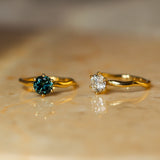 Two gold rings displayed on a beige stone surface. The left ring features a round-cut deep blue gemstone, and the right ring features a round-cut clear gemstone.
