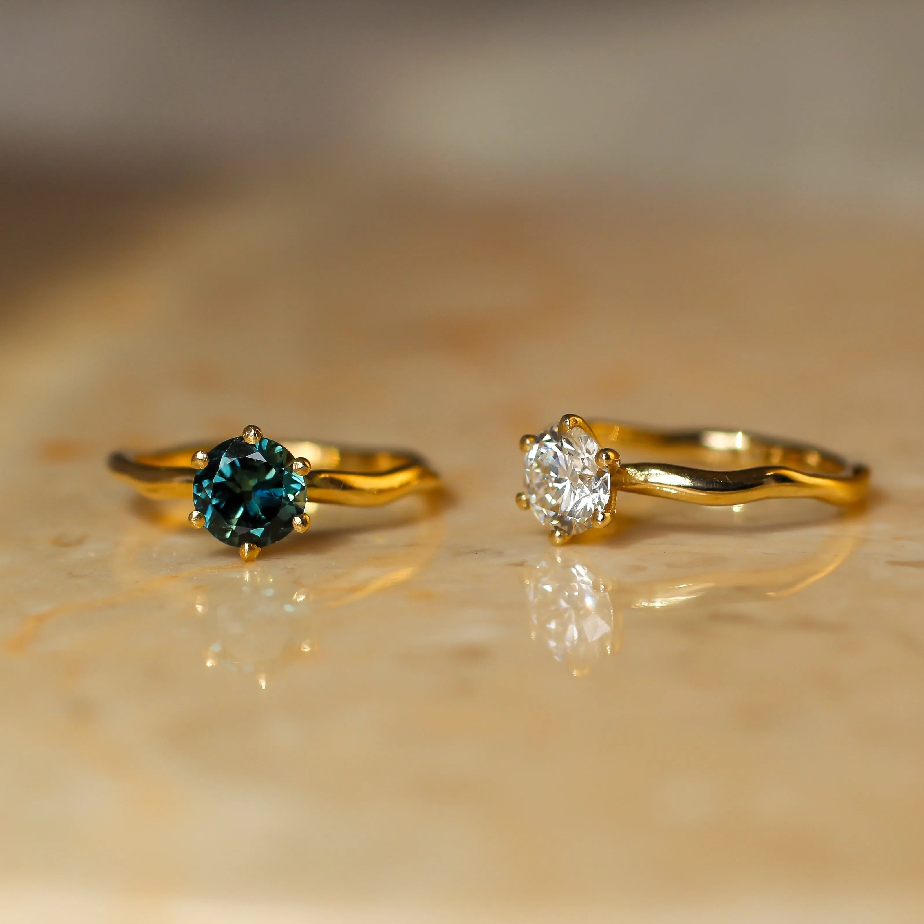 Two gold rings displayed on a beige stone surface. The left ring features a round-cut deep blue gemstone, and the right ring features a round-cut clear gemstone.