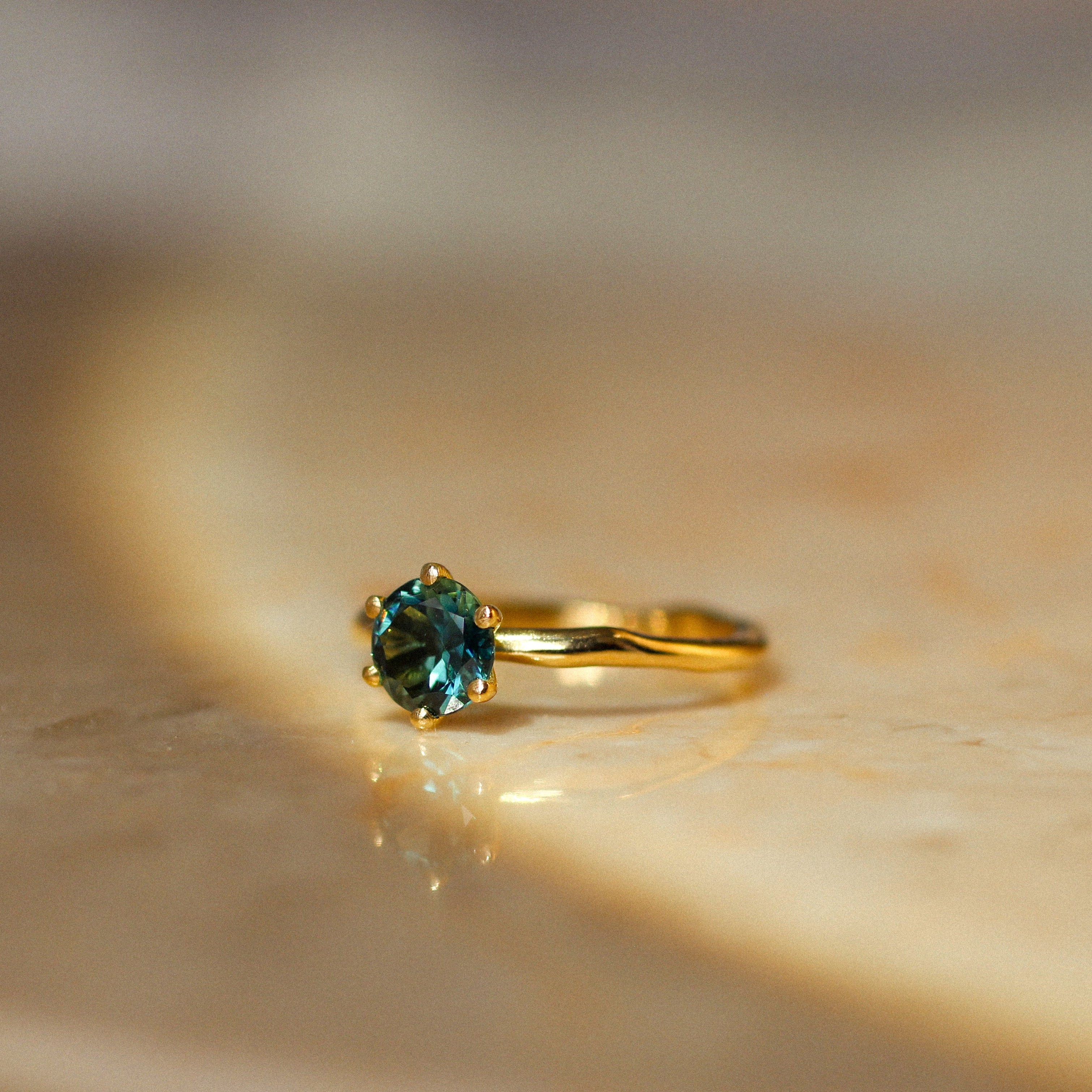 A gold ring with a round-cut blue-green gemstone and organic band, resting on a textured beige stone surface.