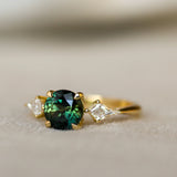 A yellow gold ring featuring a round-cut dark green gemstone at the center, flanked by two small clear kit side diamonds, photographed on a soft beige textured surface.