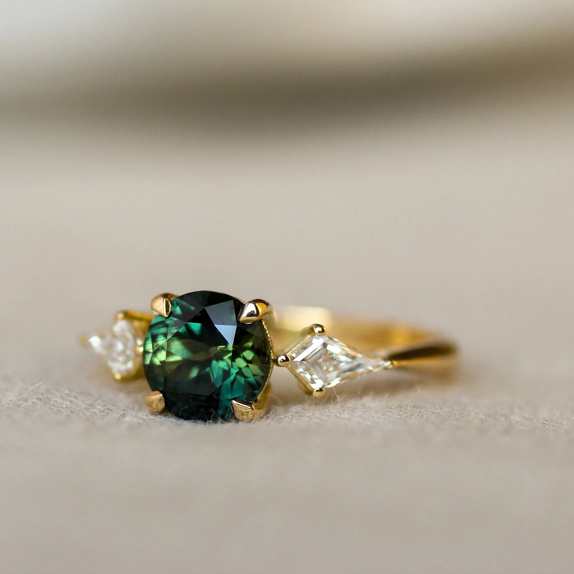 A yellow gold ring featuring a round-cut dark green gemstone at the center, flanked by two small clear kit side diamonds, photographed on a soft beige textured surface.