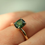 close up of hand wearing cushion cut green sapphire solitaire engagement ring with textured gold band