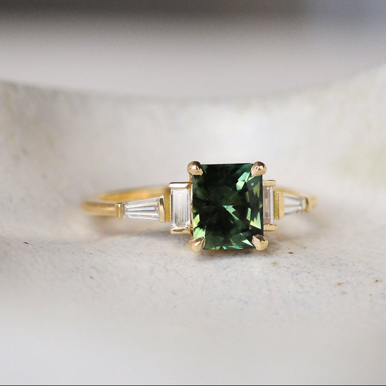 Art deco style engagement ring with a green gemstone and baguette cut diamonds on a white surface