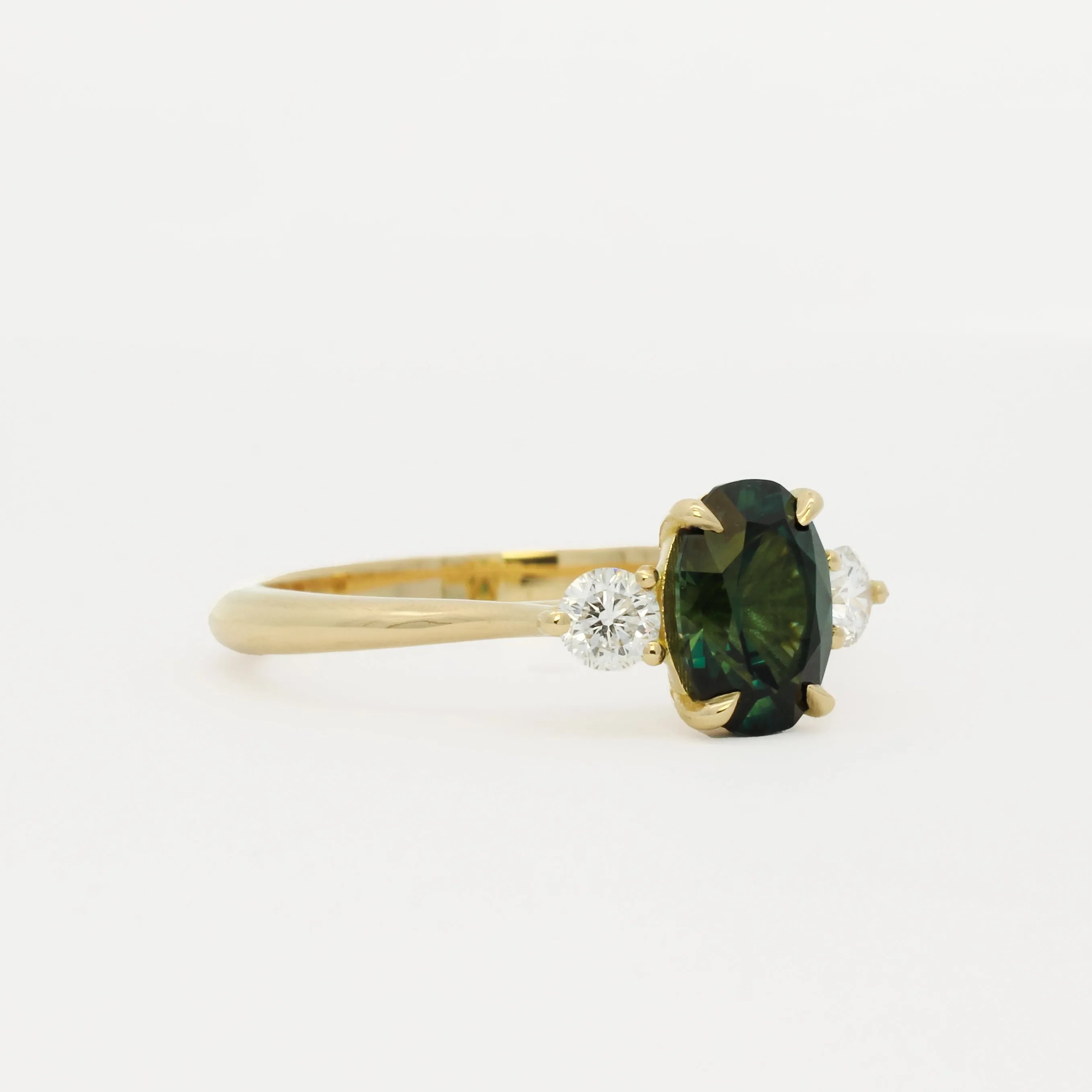 side view of a 18ct gold engagement ring with round diamonds and oval blue/green sapphire on a white background