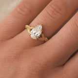 gold ring with a pear-shaped diamond and leaf detail on a hand against a neutral background