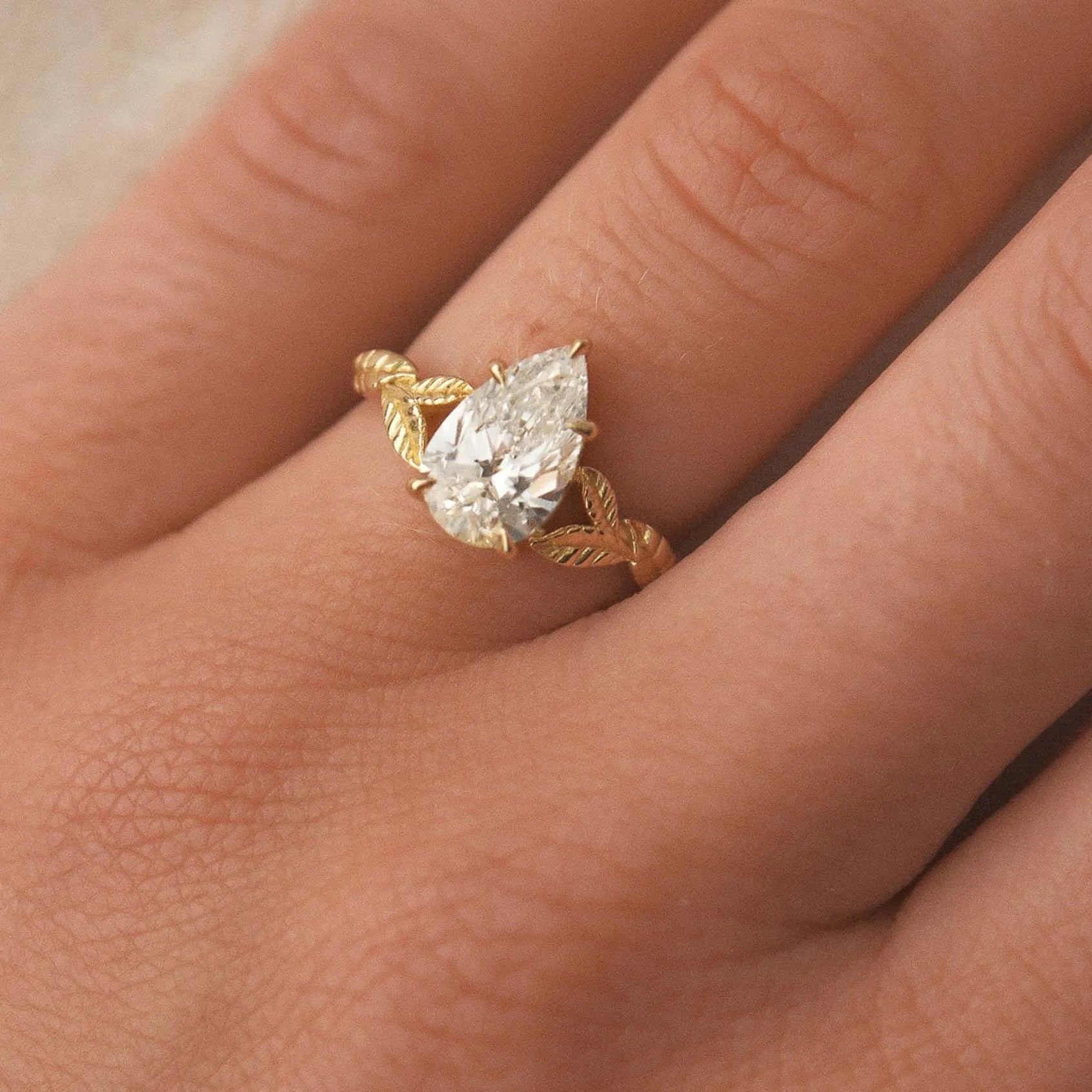 gold ring with a pear-shaped diamond and leaf detail on a hand against a neutral background