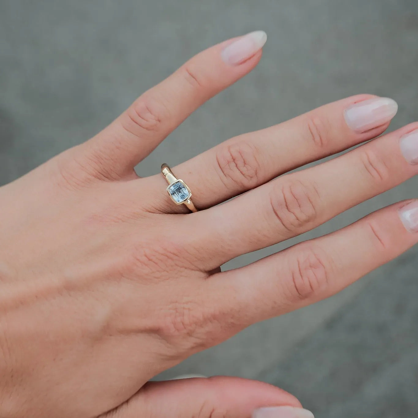 A gold ring with a small light blue gemstone worn on a person's ring finger, shown against a soft gray textured background.