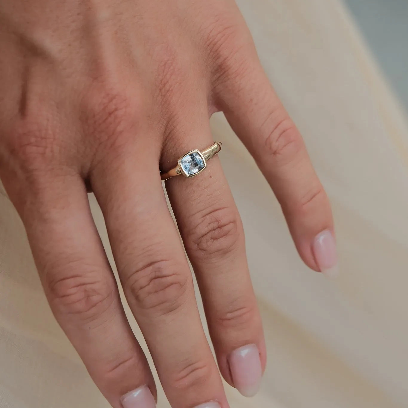 A close-up of a hand wearing a gold ring with a rectangular light blue sapphire, resting against a soft beige fabric background.