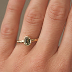 Gold engagement ring with a green gemstone and hammered texture on a person's finger against a neutral background