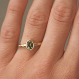 Gold engagement ring with a green gemstone and hammered texture on a person's finger against a neutral background