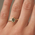 Gold engagement ring with a green gemstone and hammered texture on a person's finger against a neutral background