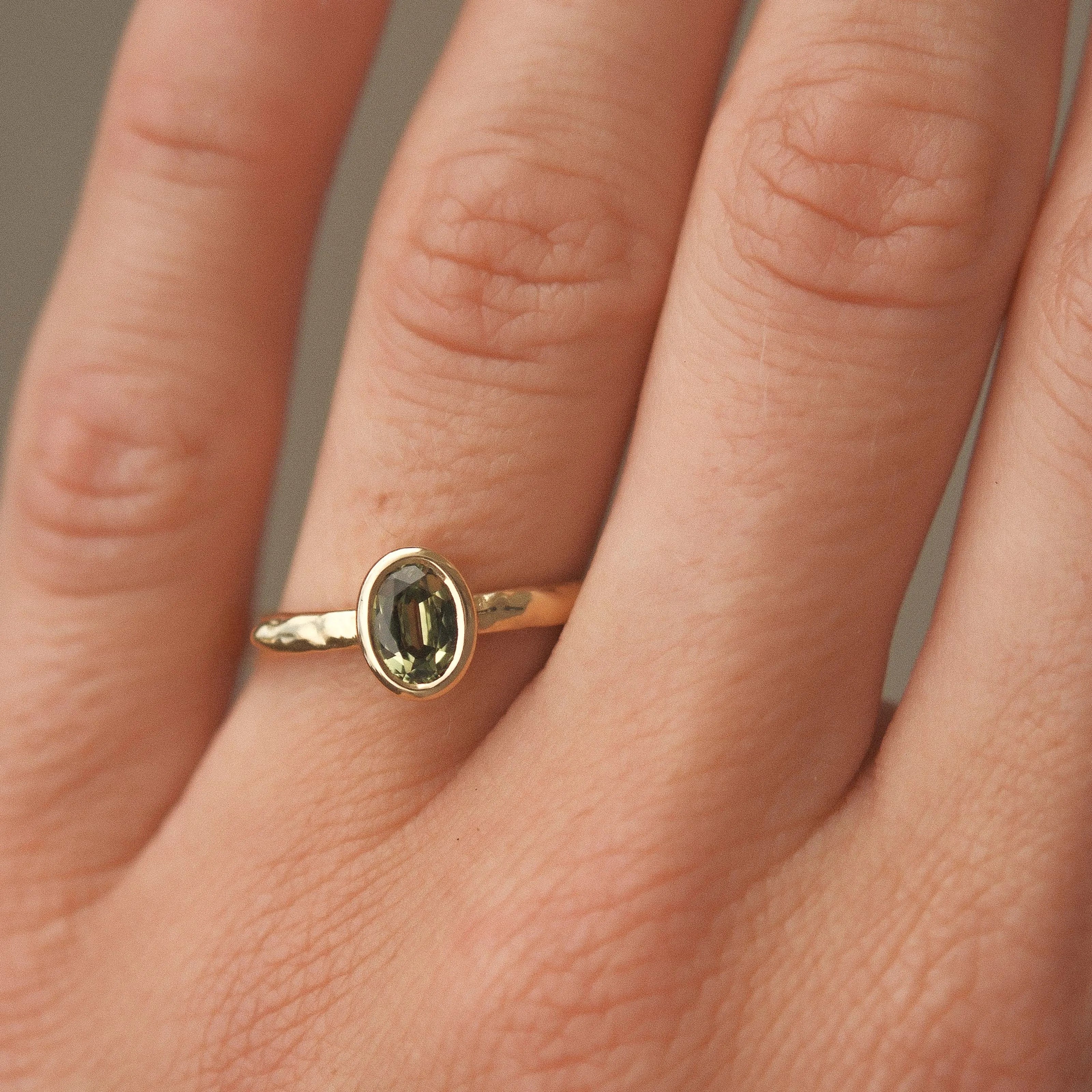 Gold engagement ring with a green gemstone and hammered texture on a person's finger against a neutral background