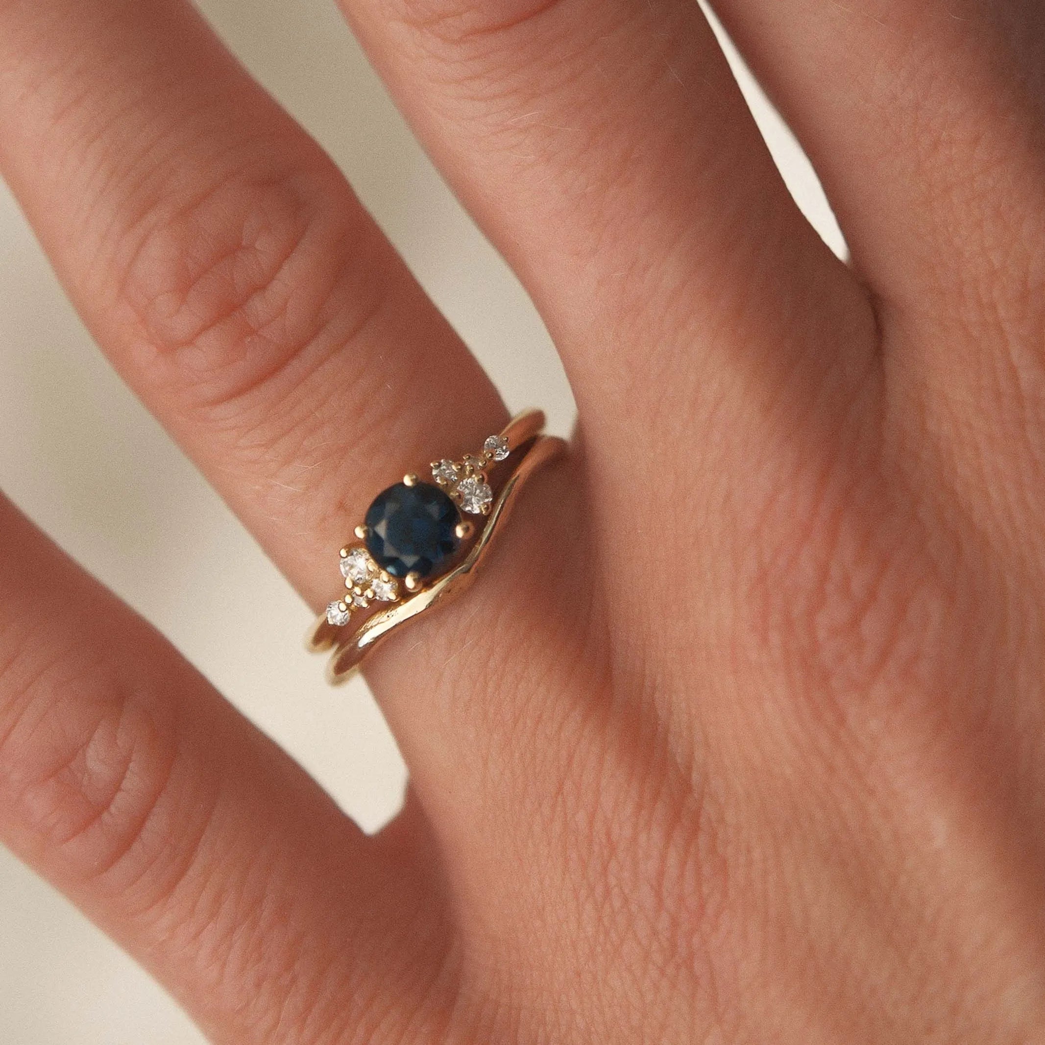 Blue sapphire engagement ring with diamonds worn with a slim soft curve wedding ring on a hand against a neutral background