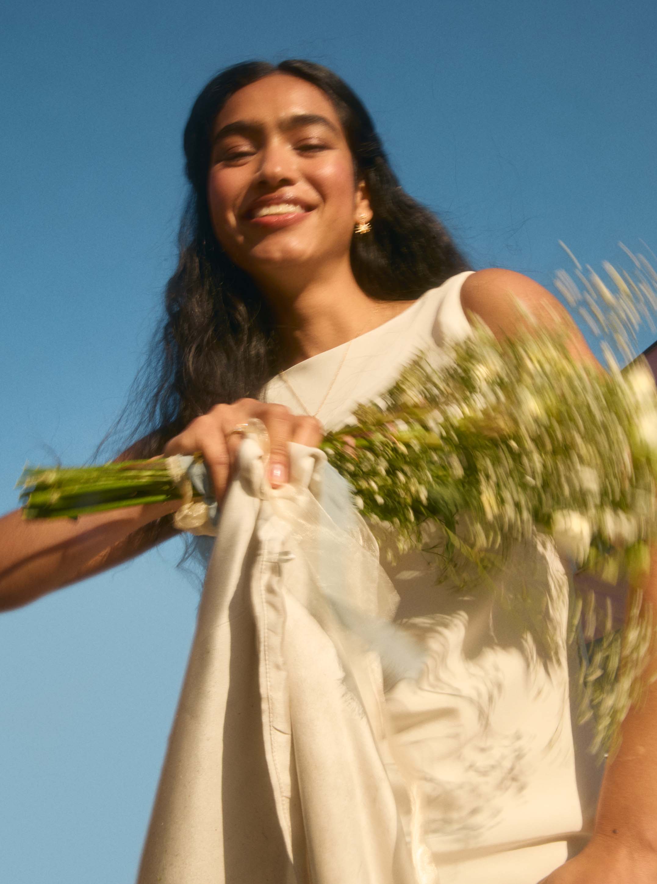 bridal model wearing dangle earrings, moving a bouquet so it blurs and smiling to camera against a blue sky