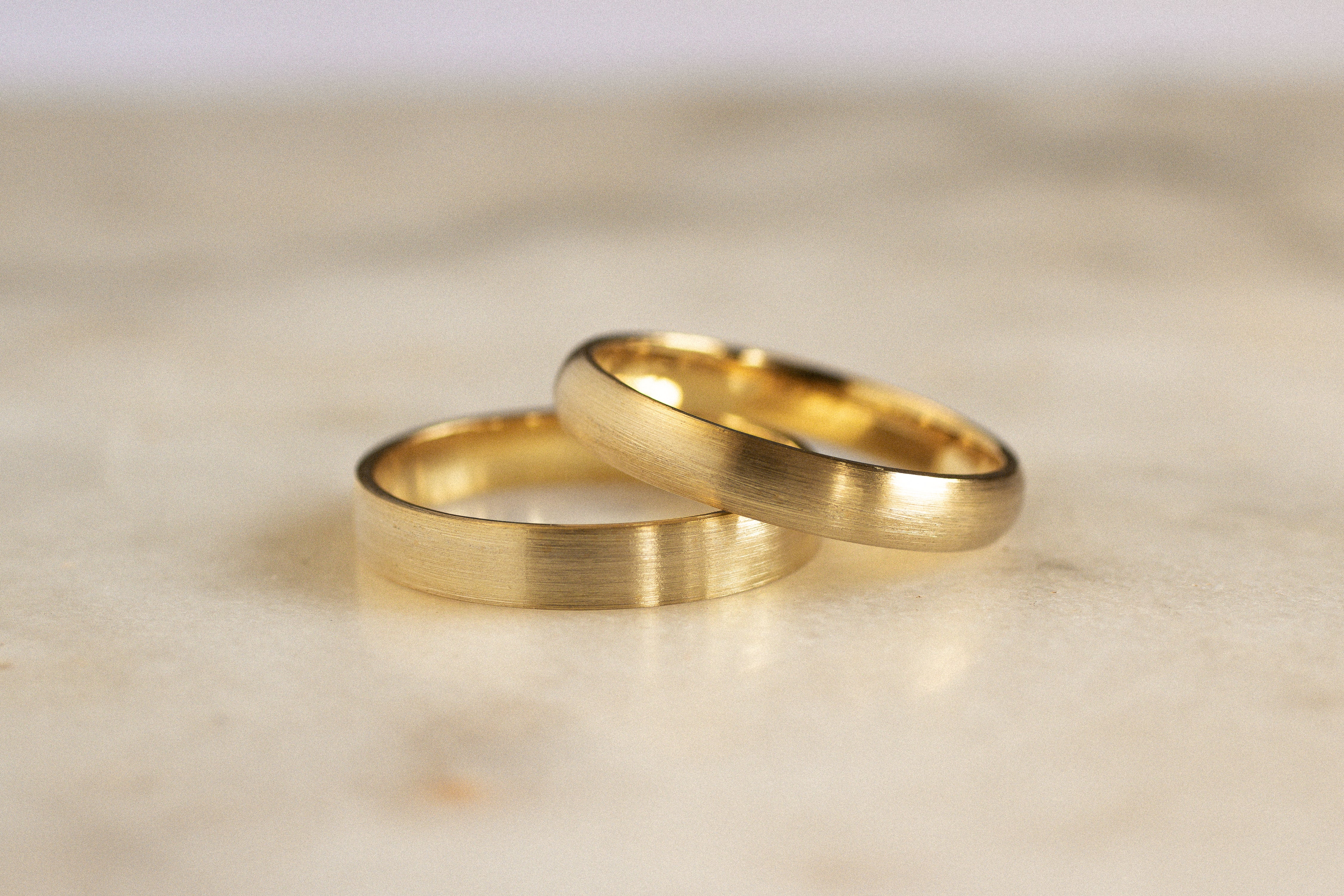 two gold wedding rings with brushed finish in flat and d shapes leaning on one another on marble surface
