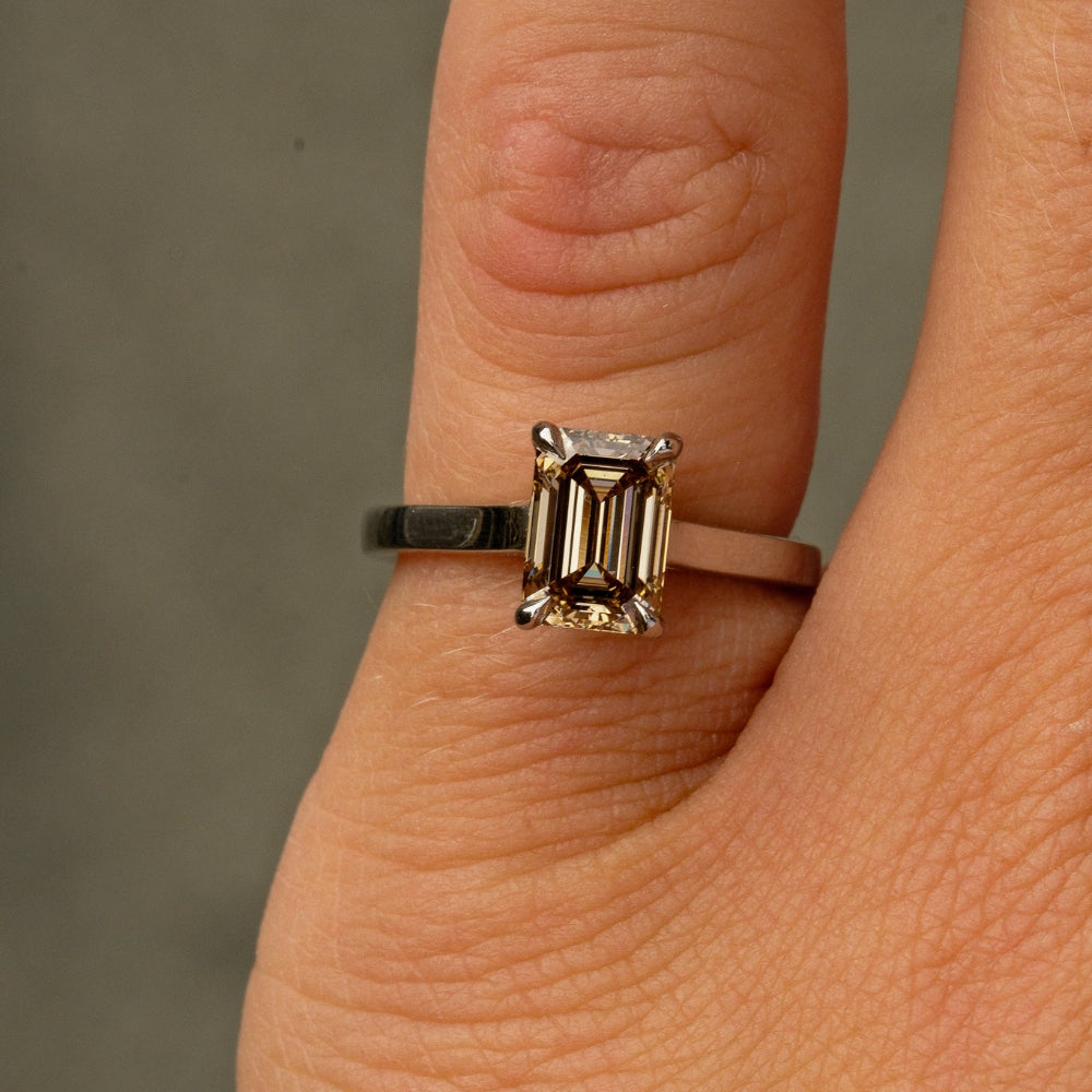 A close-up of a ring worn on a finger, featuring an emerald-cut clear brown diamond in a silver-toned setting with a dark band.
