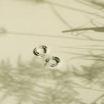 A pair of small silver hoop earrings resting on a warm beige surface with soft plant shadows cast across it.