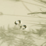 A pair of small silver hoop earrings resting on a warm beige surface with soft plant shadows cast across it.