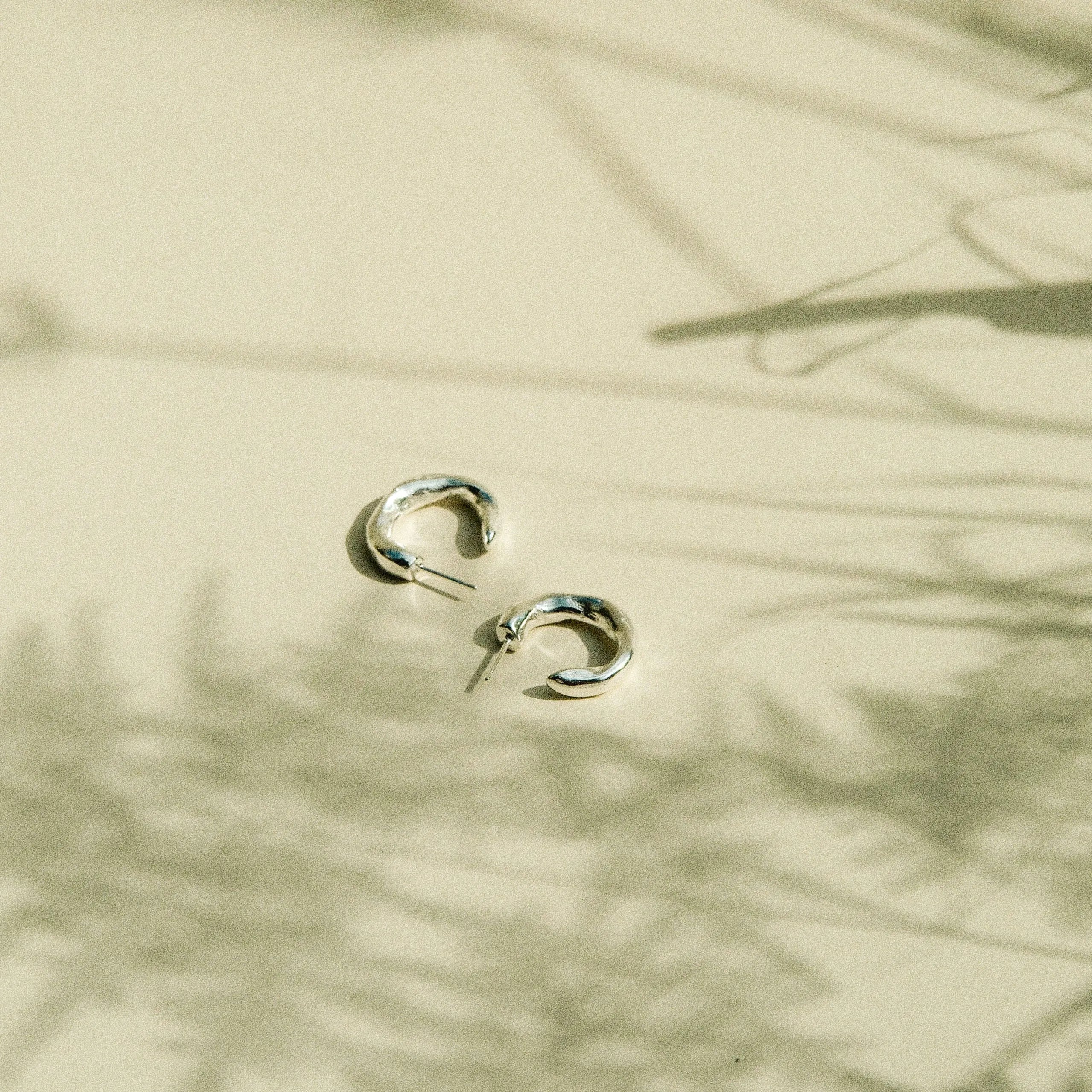 A pair of small silver hoop earrings resting on a warm beige surface with soft plant shadows cast across it.