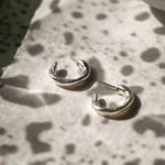 A pair of polished silver hoop earrings resting on a textured white surface with a mottled dark green background and a ceramic bowl in the upper right.