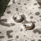 A pair of polished silver hoop earrings resting on a textured white surface with a mottled dark green background and a ceramic bowl in the upper right.